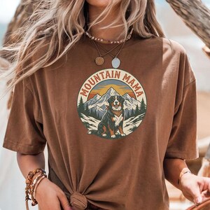 May include: A brown t-shirt with a circular graphic. The graphic features a dog, mountains, and the text "MOUNTAIN MAMA". The design uses a vintage style with brown, orange, and green colours. The t-shirt has short sleeves and a relaxed fit.