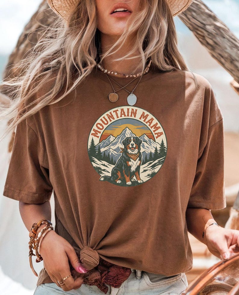 May include: A brown t-shirt with a circular graphic. The graphic features a dog, mountains, and the text "MOUNTAIN MAMA". The design uses a vintage style with brown, orange, and green colours. The t-shirt has short sleeves and a relaxed fit.