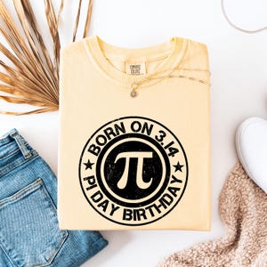 Może przedstawiać: Jasnożółta bluza z czarną grafiką z napisem "BORN ON 3.14 PI DAY BIRTHDAY" wokół symbolu pi. Koszulka jest złożona, z niebieskimi dżinsami i naszyjnikiem.
