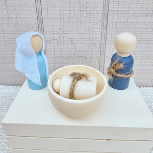 May include: A wooden nativity scene featuring three figures: a blue-robed Mary figure with a white head covering, a blue-robed Joseph figure, and a baby in a wooden bowl. The figures are on a white wooden box.