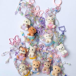 May include: A collection of keychains featuring small animal figurines, including dogs, cats, and bears, each wearing a pastel-colored dress. The keychains are adorned with beaded strands and decorative charms in shades of pink, purple, and blue.