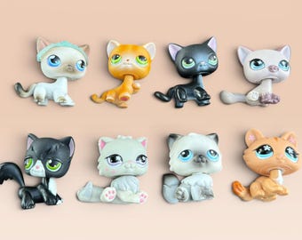 May include: A collection of eight small plastic cat figurines with oversized heads and large eyes. The cats feature various colors including white, orange, black, and grey, with some displaying patterned markings, painted facial details, and small paw prints.