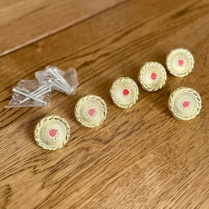 May include: Six gold cabinet knobs with a red centre and a rope-like border are arranged on a wooden surface. A small bag of screws is also present. These decorative knobs are likely for furniture or home decor.