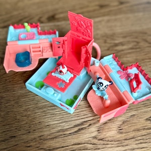 May include: Miniature playsets in coral and light blue. One set is open, revealing a small figure with a red and white helmet. Another set features a panda figure with a blue tutu. The sets are on a wooden surface.