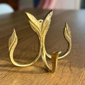 May include: A gold-coloured metal wall hook with a leaf design. The hook has three curved leaf-shaped arms and a small hook at the bottom. The hook is designed to be mounted on a wall.