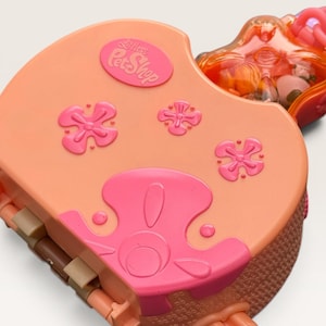 May include: A peach-colored Littlest Pet Shop toy case featuring a pink embossed logo, three raised flower motifs, and a bright pink decorative overlay. The case includes a translucent orange compartment at the top with visible contents inside.