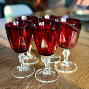 May include: A set of six ruby red wine glasses with clear, decorative stems and circular bases. The glasses feature a faceted design, reflecting light. The glasses are arranged on a wooden surface, creating a vintage and elegant aesthetic.