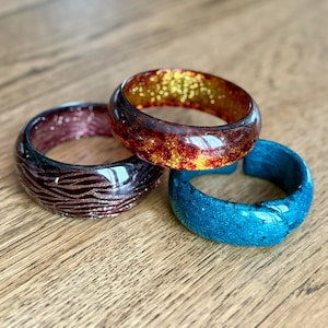Peut inclure: Trois bracelets en résine rouges, marron et bleus. L'un est rouge foncé avec des paillettes dorées, un autre est rouge foncé avec un motif zébré et le troisième est bleu vif avec des paillettes.