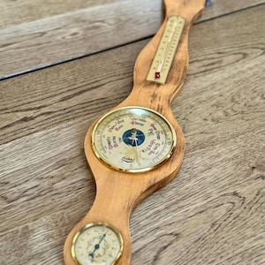 May include: A wooden weather station with a thermometer, barometer, and hygrometer. The barometer has a gold-colored bezel and a dial indicating weather conditions. The thermometer displays the temperature in Fahrenheit. The hygrometer is at the bottom.