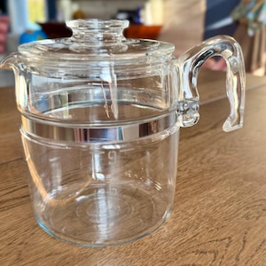 May include: A clear glass coffee pot with a lid and handle. The pot has a silver band around the middle and measurement markings on the side. The handle is curved and attached to the side of the pot. The lid has a knob on top.