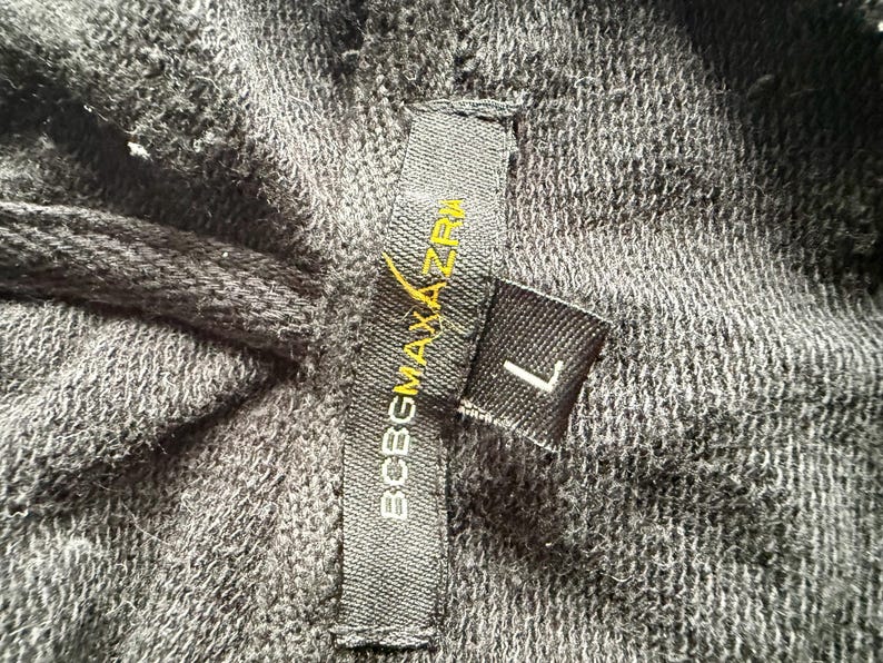 May include: Close-up of dark gray fabric with a tag that reads "BCBGMAXAZRIA" in white and yellow lettering. A smaller tag indicates the size "L". The fabric appears to be a knit or woven material.