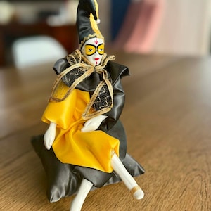 May include: A porcelain jester figurine with a black and yellow hat and outfit, and a white face with painted features. The jester is seated on a grey cushion, with gold accents on the outfit and feet. The figurine is a decorative item.