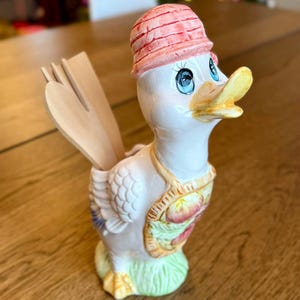 May include: A ceramic duck-shaped utensil holder with a red hat and a yellow beak. The duck has blue eyes and a decorative apron with floral details. Wooden utensils are placed inside the holder. The holder is on a wooden surface.