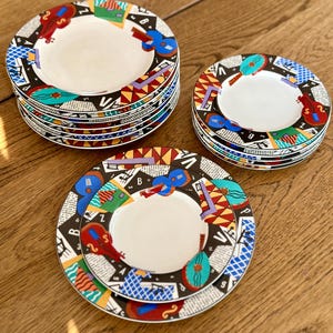 May include: A set of decorative ceramic plates with a musical theme. The plates feature a white center and a colorful border with musical instruments like guitars and violins. The border also includes geometric shapes and text elements in a black and white background.