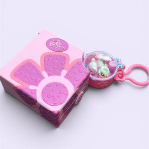 May include: A pink square Littlest Pet Shop case featuring a large purple flower graphic on the lid, paired with a small circular clear plastic dome keychain containing two miniature animal figures attached by a pink plastic ring.