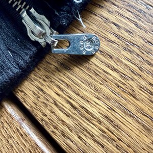 May include: Close-up of a silver zipper pull and zipper teeth on a black fabric. The zipper pull has a circular design with text. The fabric is a dark, textured material. The background is a wooden surface.