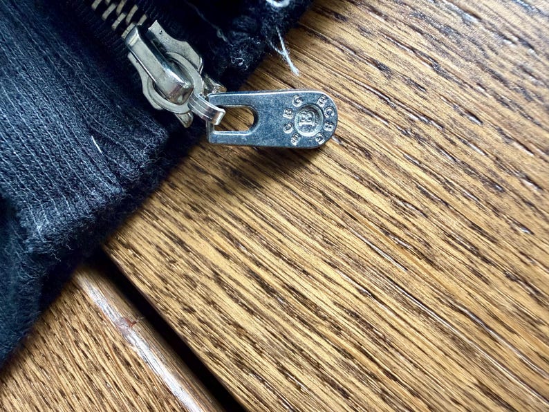 May include: Close-up of a silver zipper pull and zipper teeth on a black fabric. The zipper pull has a circular design with text. The fabric is a dark, textured material. The background is a wooden surface.