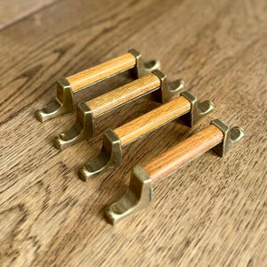 May include: Four cabinet pulls with a brass finish and light brown wooden handles. The pulls are rectangular with a curved base and are arranged diagonally on a wooden surface.