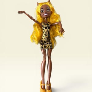 May include: A doll with dark skin, pointed ears, and long, wavy yellow hair. The doll wears a sleeveless black dress with a gold geometric pattern and matching gold Mary Jane shoes. The arms are posed in an upward, outstretched position.