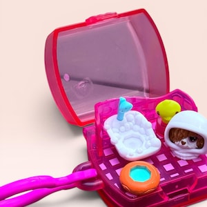 May include: A translucent hot pink plastic carrying case opens to reveal a miniature bathroom playset. Inside are a white bathtub with a blue faucet, a small yellow duck, a brown and white puppy figure wearing a white hood, and an orange donut.