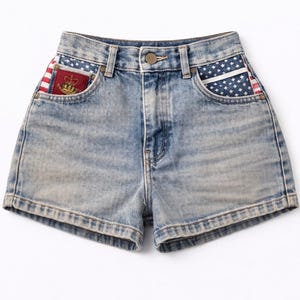 May include: Light blue denim shorts with American flag pocket accents. The pockets feature a white star design on a blue background and red and white stripes. A gold crown emblem is visible on the left pocket.