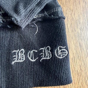 May include: Close-up of a dark gray ribbed knit fabric with the embroidered letters "BCBG" in a stylized font. The fabric has a distressed, frayed edge and visible stitching.