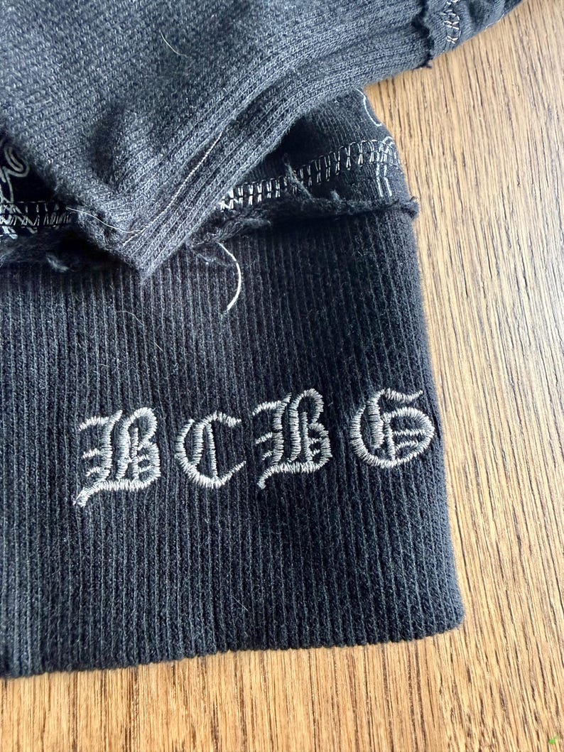 May include: Close-up of a dark gray ribbed knit fabric with the embroidered letters "BCBG" in a stylized font. The fabric has a distressed, frayed edge and visible stitching.