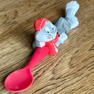 May include: A Bugs Bunny spoon featuring a red spoon bowl and handle, with Bugs Bunny's head and torso in the center. The character is wearing a red outfit and hat, with gray and white accents. The spoon is made of plastic.