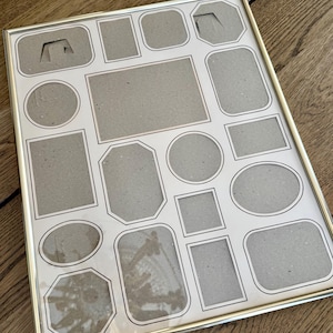 May include: A gold-coloured metal multi-photo picture frame. It features a variety of photo cutouts in shapes like rectangles, squares, circles, and ovals. The background is a light cream colour.