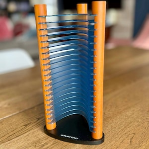 May include: An orange and blue CD storage rack. The rack has two orange vertical supports with blue plastic dividers. The base is black with the word "ATLANTIC" printed on it. The rack is designed to hold multiple CDs.
