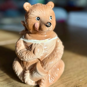 May include: A ceramic bear-shaped honey pot in shades of brown and tan. The bear is depicted sitting, holding a honey pot with the word "HONEY" on it. The bear has a pink tongue sticking out and a white collar.