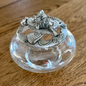 May include: A clear glass trinket dish with a decorative pewter lid. The lid features a nautical-themed design with a figure, a map, and rope detailing. The dish is round and sits on a wooden surface.