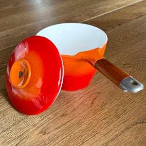 Peut inclure: Une casserole émaillée orange et rouge avec une poignée en bois et un couvercle assorti. L'intérieur de la casserole est blanc. Le couvercle est sur le côté, révélant l'intérieur de la casserole. La poignée est en bois et a une extrémité en métal.