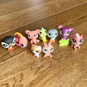 May include: A collection of colorful, small plastic animal figurines, including a penguin, fish, pig, bird, koala, and rabbit. The toys are in various shades of pink, yellow, gray, and purple, with detailed features and large eyes. They are arranged on a wooden surface.