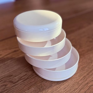 May include: A stack of four off-white, round, plastic containers. The top container has a lid, and the other three are open bowls. The containers are stacked on top of each other, with the bottom container resting on a wooden surface.