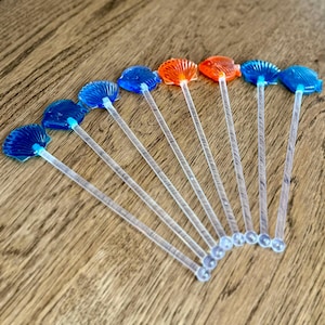 May include: A collection of seashell-shaped cocktail stirrers. The stirrers have clear stems and colorful tops in shades of blue and orange. The seashell tops are made of a translucent material. The stirrers are arranged on a wooden surface.