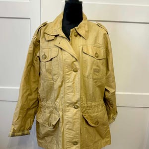 May include: Tan jacket with multiple pockets, button closures, and a drawstring waist. The jacket has a collared neckline, epaulettes, and a zipper closure. The sleeves have a plaid lining. The jacket is displayed on a black mannequin.