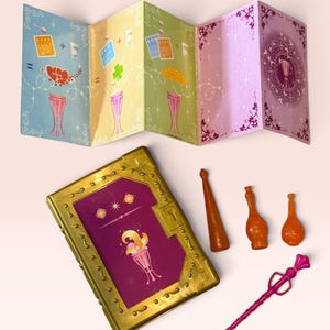 May include: A gold-colored book with a purple cover featuring a graphic of a goblet, a pink plastic wand, three small orange plastic bottles, and a multi-colored accordion-style paper instruction sheet with illustrations of goblets and symbols.