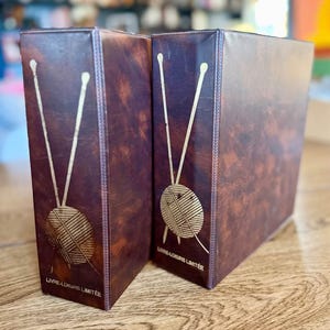 May include: Two brown leather-bound boxes with a gold-coloured knitting needle and yarn ball design. The boxes have the text "LIVRE-LOISIRS LIMITEE" at the bottom. The boxes are standing upright.