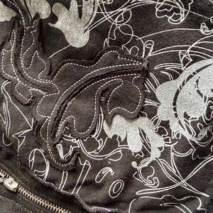 May include: Close-up of a black fabric with a floral pattern in white and gray. A leaf-shaped appliqu&eacute; with white stitching is visible. A silver zipper is partially visible on the left side of the image.