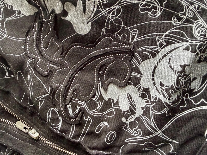 May include: Close-up of a black fabric with a floral pattern in white and gray. A leaf-shaped appliqu&eacute; with white stitching is visible. A silver zipper is partially visible on the left side of the image.