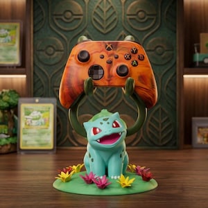 Puede incluir: Una figura de Bulbasaur turquesa sosteniendo un mando de Xbox naranja y marrón. La figura está sobre una base verde con flores coloridas. El fondo incluye cartas de Pokémon enmarcadas y una estantería de madera.