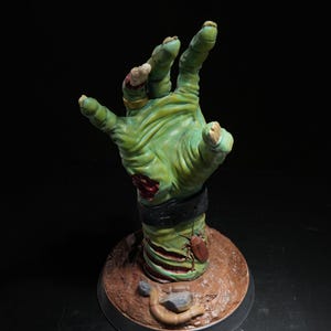 Könnte beinhalten: Eine grüne, zombieartige Handskulptur mit offenen Wunden und einem schwarzen Armband. Die Hand ragt aus einer braunen, strukturierten Basis empor. Ein großes Insekt ist am Arm befestigt. Eine einzigartige Halloween-Dekoration.