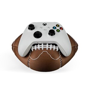May include: A white Xbox controller rests on top of a brown football. The controller has black joysticks and buttons with colored X, Y, A, and B buttons. The football is brown with white stitching.