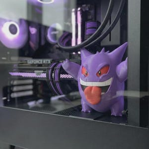 May include: A purple Gengar figurine with a red tongue and eyes stands inside a computer case. The computer case has a glass side panel, revealing internal components and purple lighting. The graphics card reads "GEFORCE RTX".
