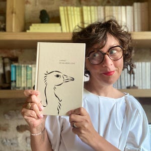Peut inclure: Un carnet beige avec le texte "CARNET DE MA MERE L'OYE" et un dessin au trait noir d'une tête d'oiseau. Le carnet est tenu par une personne portant des lunettes et un haut blanc, avec une bibliothèque en arrière-plan.