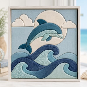 May include: A square framed fabric wall art piece featuring a layered textile design of a blue dolphin leaping over stylized ocean waves. The composition includes a large cream-colored sun and two fluffy clouds against a light blue background.