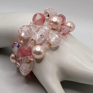 Könnte beinhalten: Ein rosa Perlenarmband mit verschiedenen Perlenformen und -größen. Das Armband hat runde, ovale und facettierte Perlen in Rosa- und Klartönen. Einige Perlen haben ein Wirbeldesign. Das Armband wird auf einer weißen Oberfläche präsentiert.