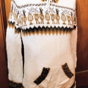 May include: White knit poncho with a hood and a front pocket. The poncho features brown and black geometric patterns around the collar, cuffs, and hem, and a repeating pattern of stylized figures across the chest. The hem has a fringe.