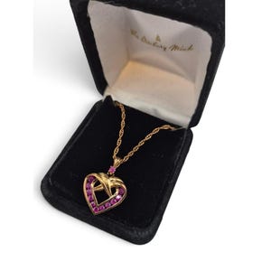 May include: A gold-tone heart-shaped pendant necklace featuring small pink stones set along the frame and a single pink stone at the top bail. The necklace is presented in a black velvet-lined box with The Danbury Mint text inside.